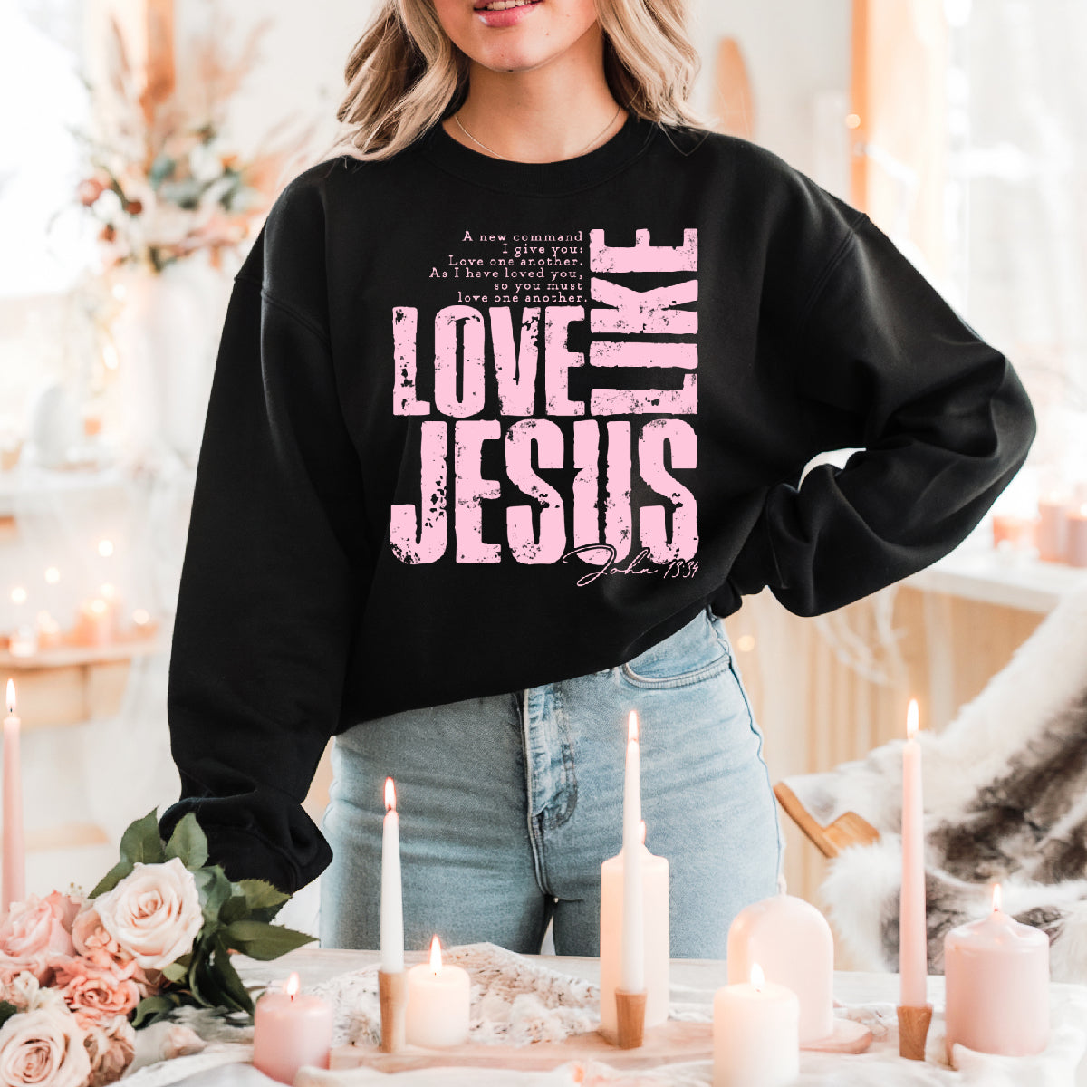 Person wearing a black sweatshirt with 'Love Like Jesus' text in pink, standing in a decorated room with candles and flowers.